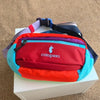 A red and blue color block waterproof nylon fanny pack with a purple strap resting on a white box.