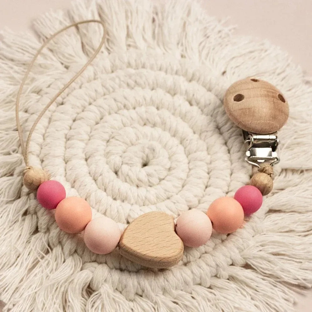 A wooden pacifier clip with crochet beads and a chain rests on a cream-colored knitted surface.