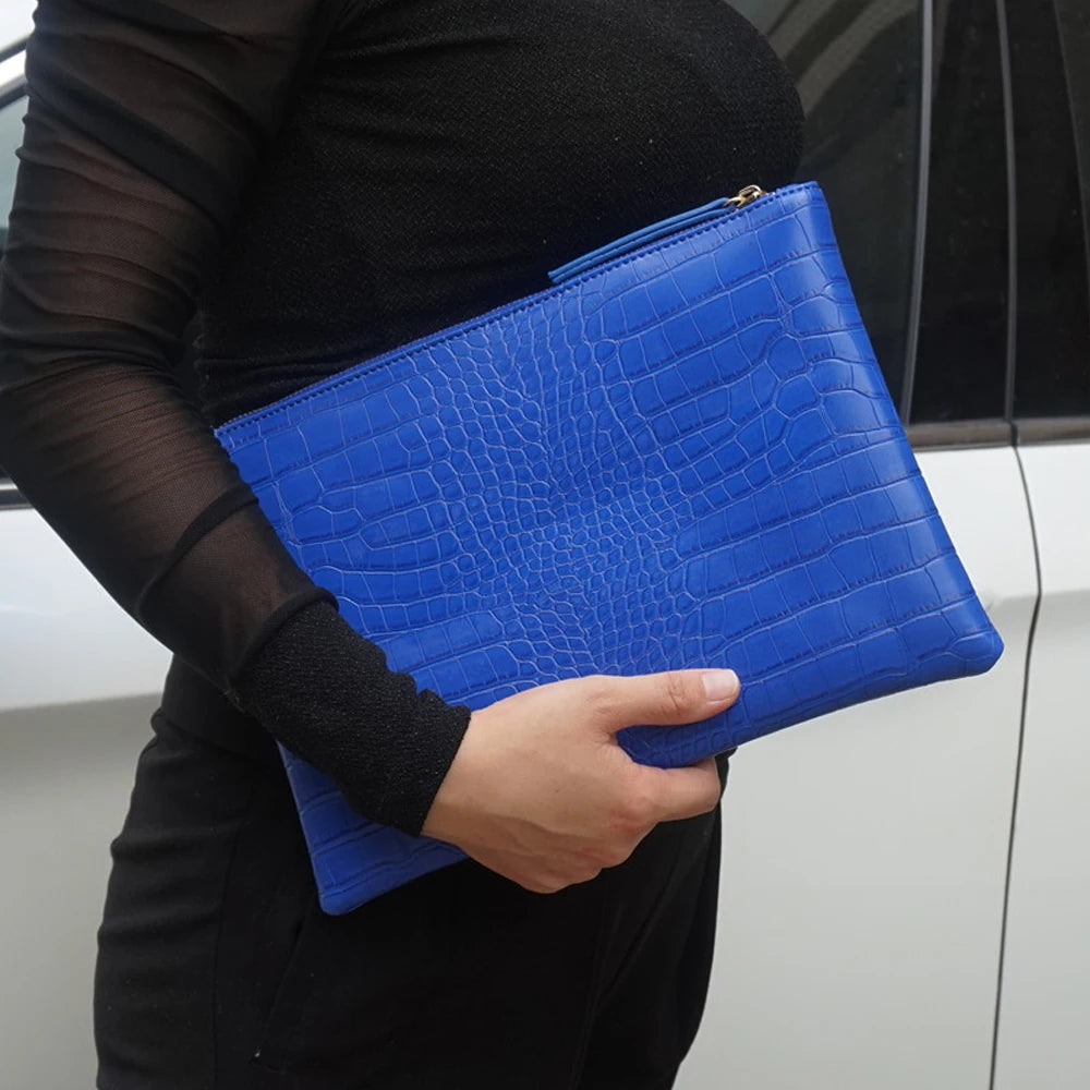 A hand holds a bright blue crocodile-embossed PU leather envelope clutch against a dark background.