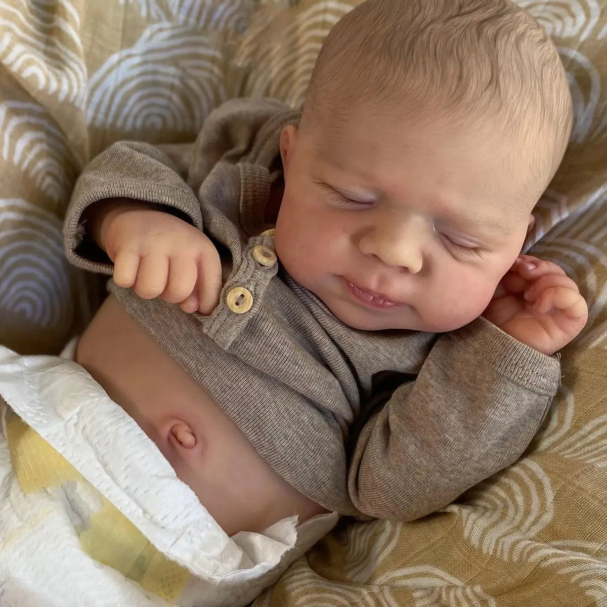 A newborn baby doll with soft skin and light hair is lying on a patterned fabric.