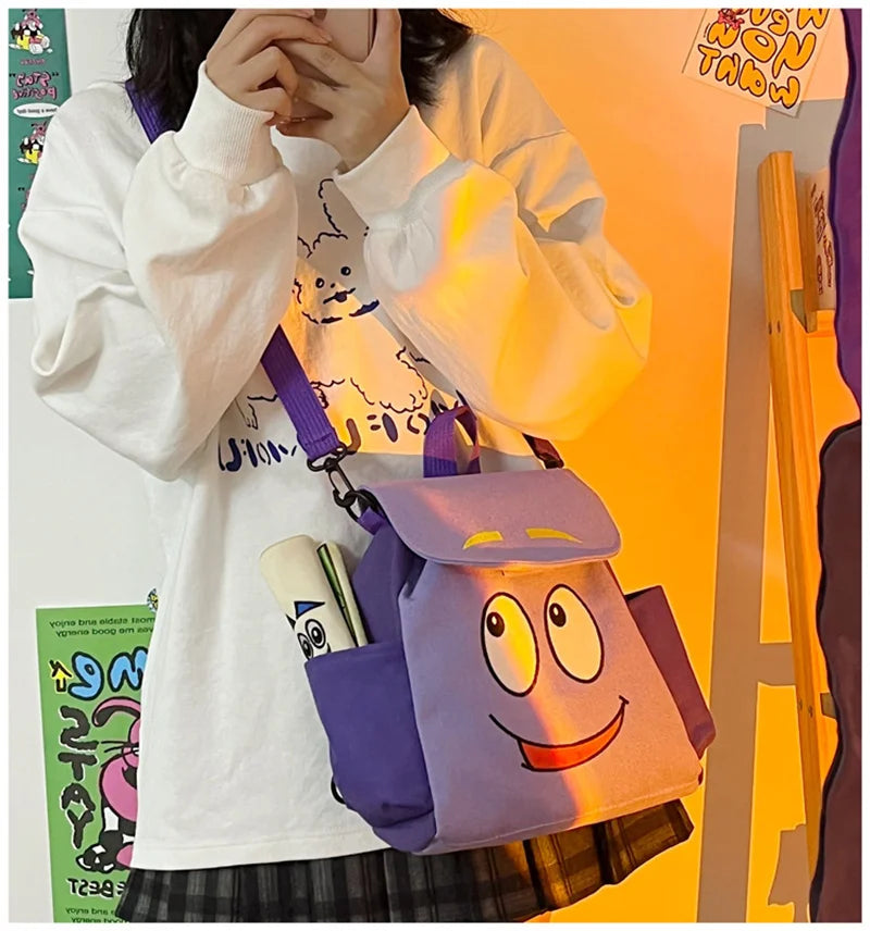 Eine Person hält einen lila und gelben Dora The Explorer Flanell-Rucksack vor einem orangefarbenen Hintergrund.