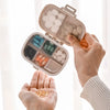 A hand opens a weekly pill organizer with eight compartments filled with white and colored pills.