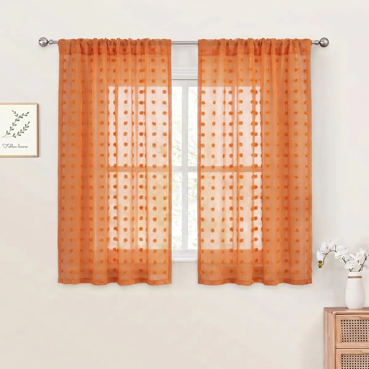 Orange sheer curtains with pom pom detailing and 3D jacquard rod pockets hanging in a window.