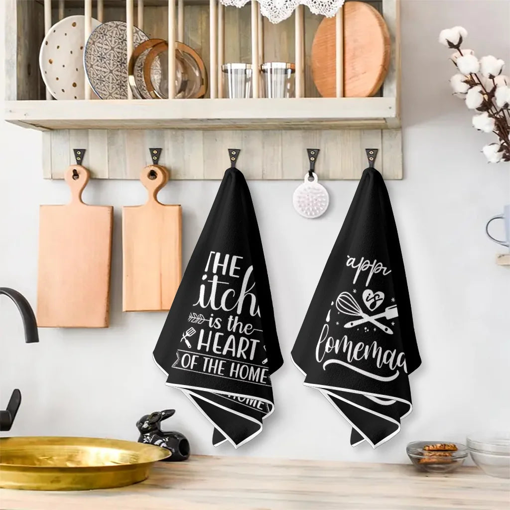 A set of two black kitchen towels featuring the slogan The itch is the heart of the home hanging on a white wall.