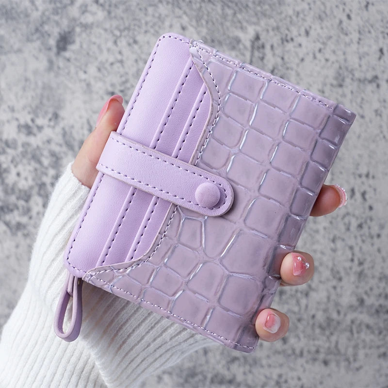 A hand holds a light purple crocodile-embossed clutch wallet with a snap closure against a gray background.