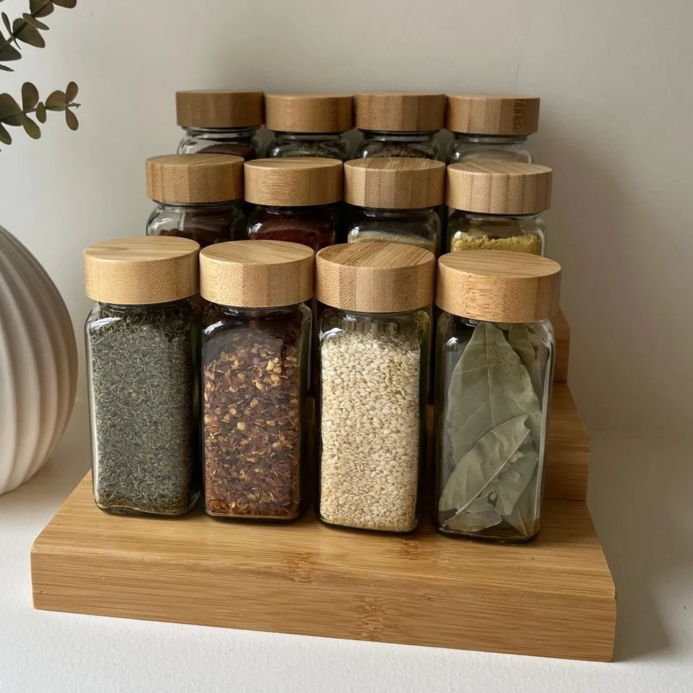 A row of clear glass spice jars with bamboo lids filled with various ground spices arranged on a wooden board.