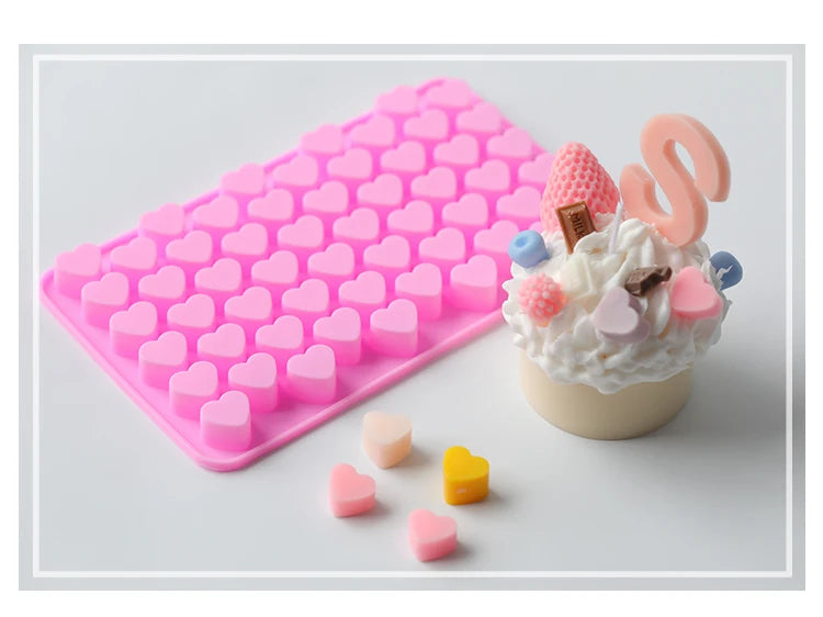 A pink silicone mold tray filled with heart-shaped cavities, next to a cupcake decoration made of pink and brown elements.