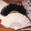 A vintage folding fan with a white lace-trimmed surface and a golden edge is displayed next to a black feather fan.
