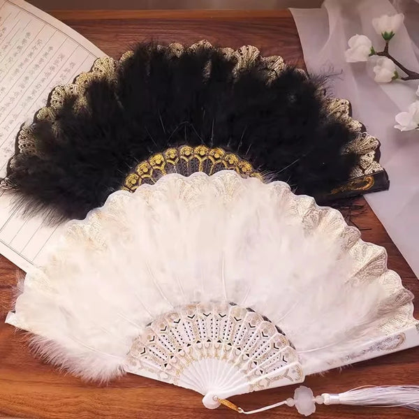 A vintage folding fan with a white lace-trimmed surface and a golden edge is displayed next to a black feather fan.