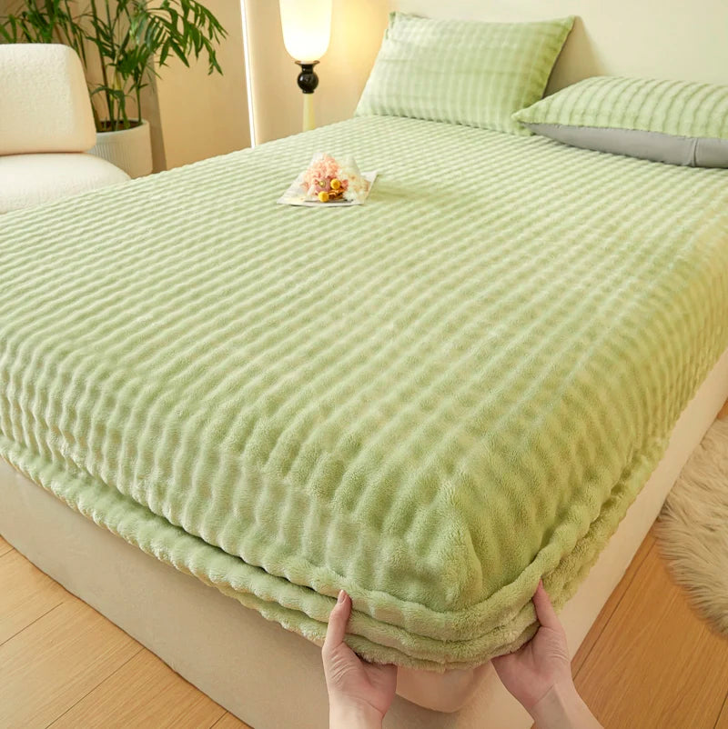 A hand is holding a light green faux fur fitted sheet on a bed.