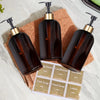 A collection of refillable gold pump soap dispenser bottles and a sheet of soap labels on a light surface.