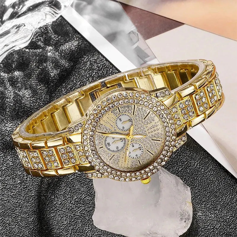 A gold rhinestone watch and jewelry set is displayed on a dark surface next to a piece of ice.