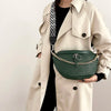 A woman wears a beige trench coat and carries a dark green woven pattern waist bag with a gold chain strap.