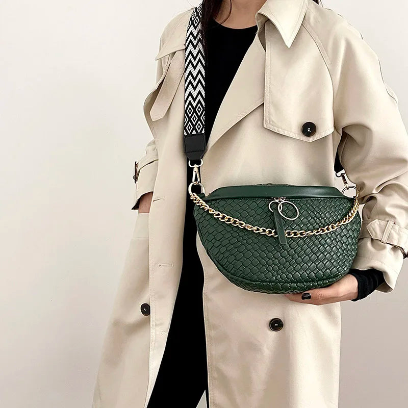 A woman wears a beige trench coat and carries a dark green woven pattern waist bag with a gold chain strap.