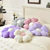 A collection of plush daisy flower throw pillows in shades of purple, pink, and gray resting on a white bed.