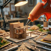 An orange hot melt glue gun is being used to apply glue to a wooden model kit on a workbench surrounded by crafting tools.