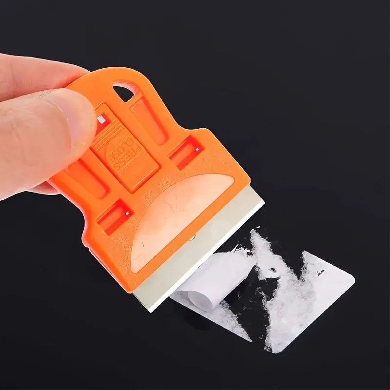 An orange compact multi-purpose scraper is shown being used to remove stickers or paint from a surface.