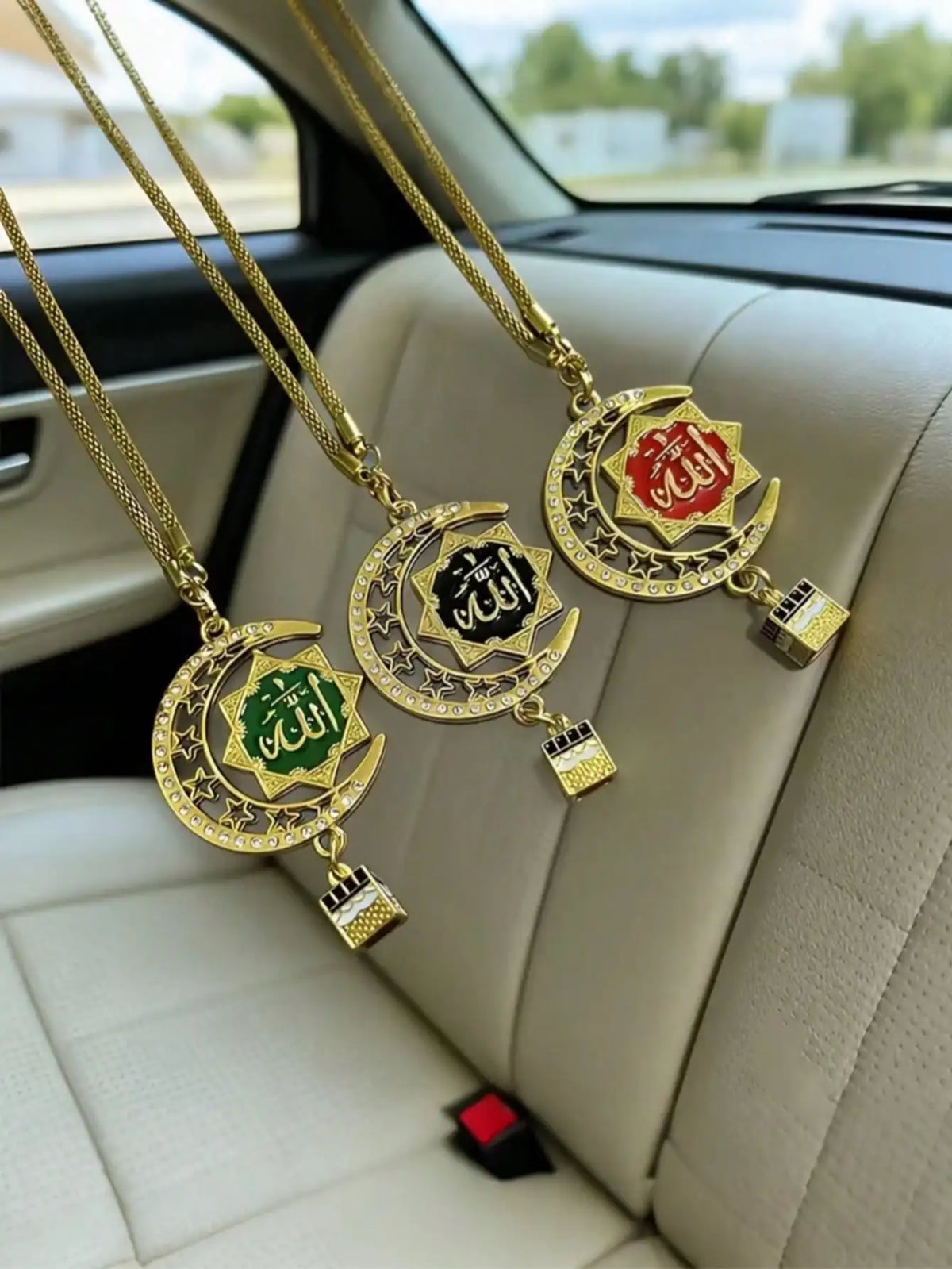 Three gold-tone Islamic calligraphy pendants on chains resting on a light-colored car seat.
