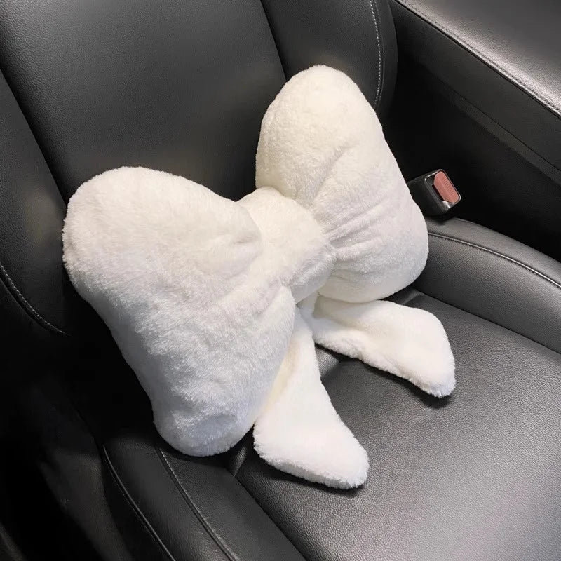 A white plush bowknot car headrest pillow with neck support resting on a dark gray car seat.