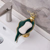 A leaf-shaped ceramic soap dish with a white soap bar and gold drain rack sits on a white bathroom counter.