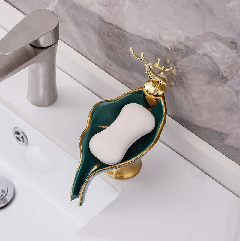 A leaf-shaped ceramic soap dish with a white soap bar and gold drain rack sits on a white bathroom counter.
