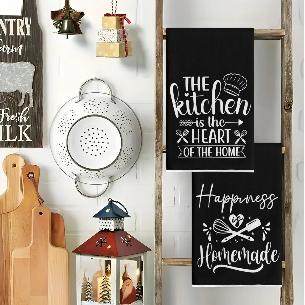 A set of two black kitchen towels featuring slogans like The Kitchen is the Heart of the Home and Happiness Homemade