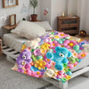 A colorful flannel throw blanket featuring various pastel-colored floral patterns and teddy bear motifs draped over a white b