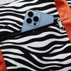 A close-up of a light blue smartphone placed inside a black and white zebra print duffle bag with an orange strap.
