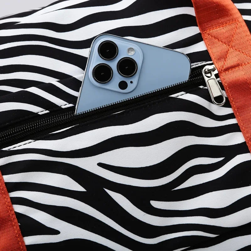 A close-up of a light blue smartphone placed inside a black and white zebra print duffle bag with an orange strap.