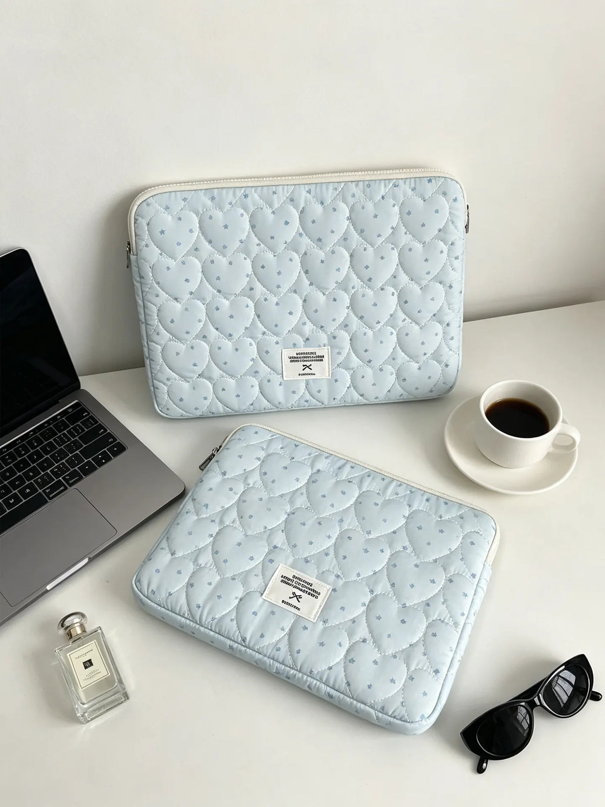 A light blue quilted laptop sleeve with a soft lining resting on a white surface next to a silver laptop and sunglasses.