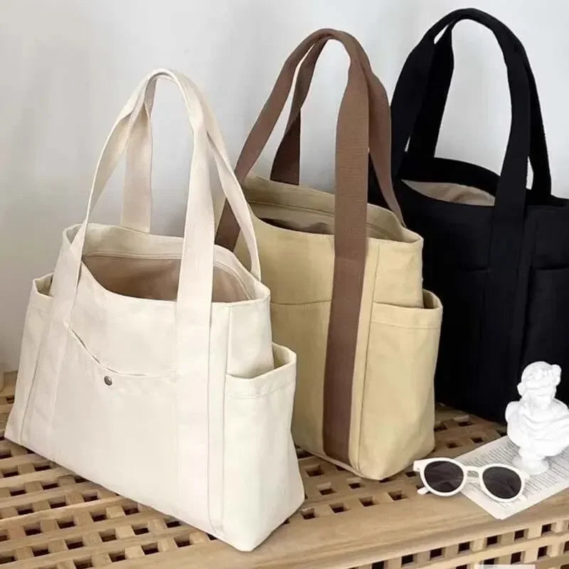 Tres bolsas de tela en lienzo en crema, beige y negro colgadas en una superficie de madera.