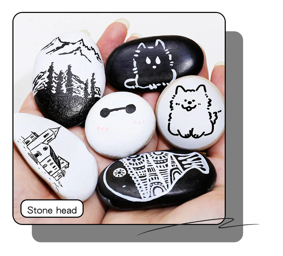 A hand holds several smooth, black and white stone head-shaped acrylic paint marker pens with various animal and architectura
