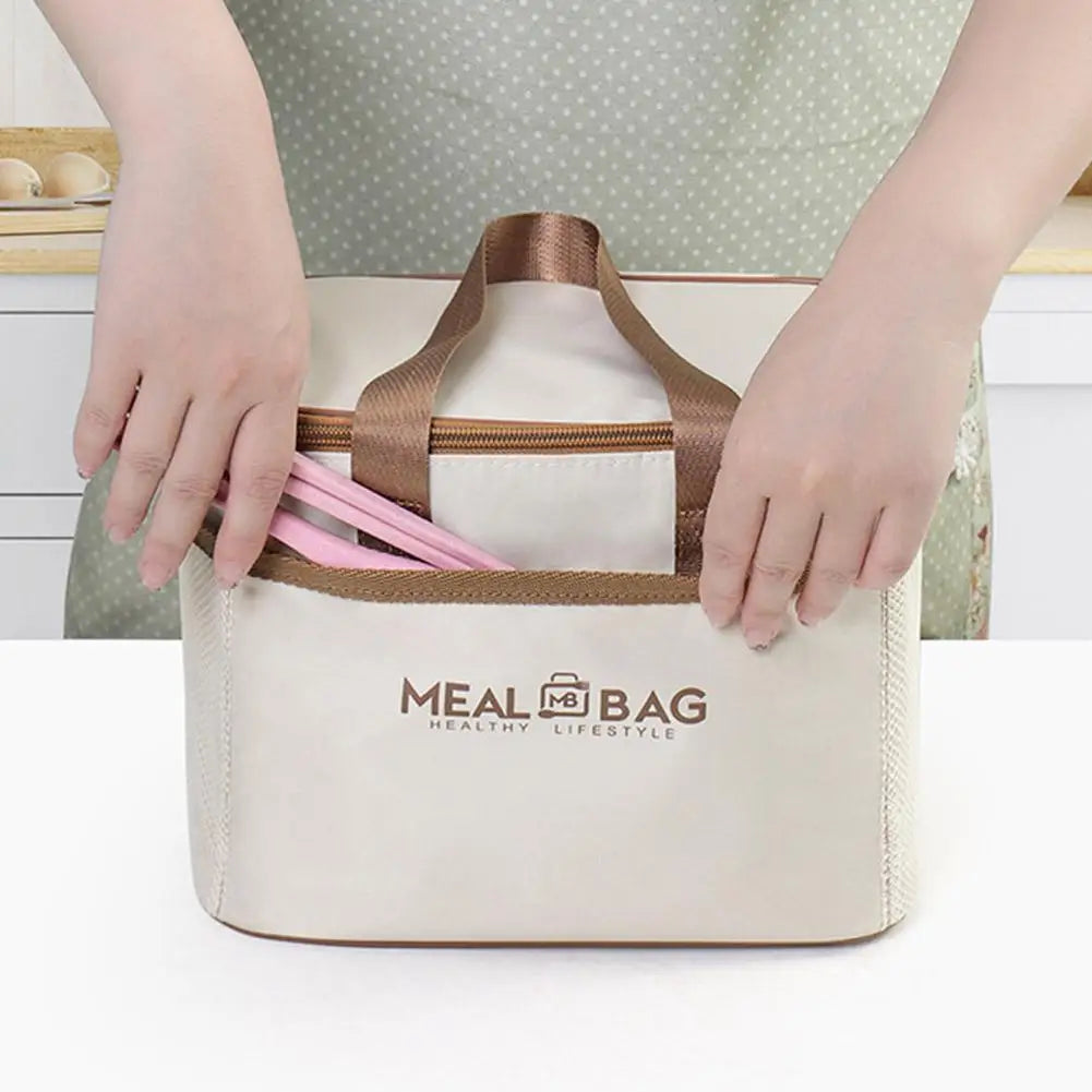 A person holds a beige insulated lunch bag with a brown strap and pink utensils on a white table.
