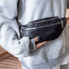 A person is holding a black nylon waist pack with a zipper on their gray sweatshirt.