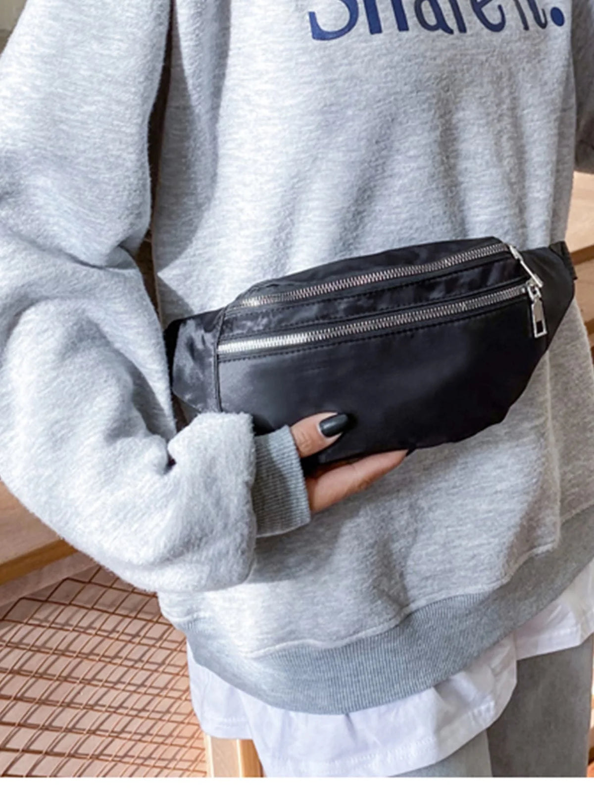 A person is holding a black nylon waist pack with a zipper on their gray sweatshirt.