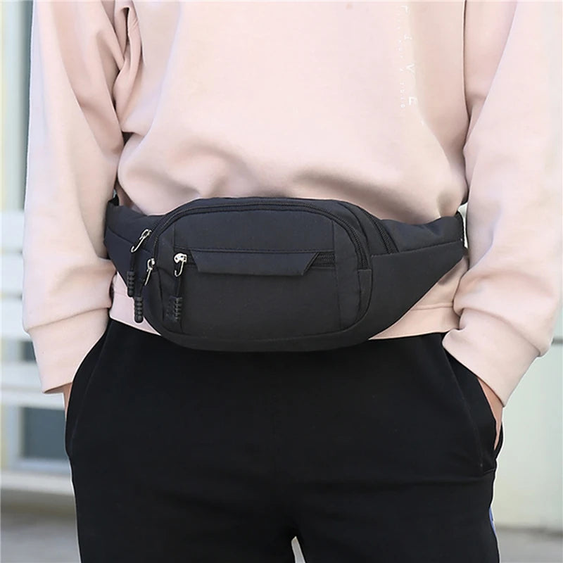 A black canvas waist pack with multiple pockets is being held by a person wearing a light pink top and black pants.