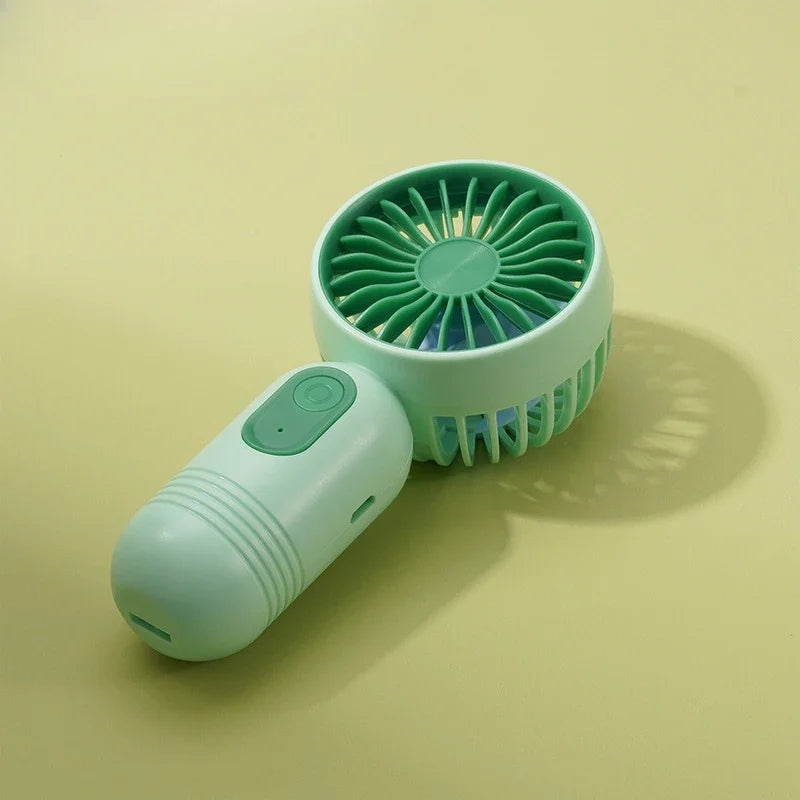 A light mint green handheld fan with a circular grille and a smooth body is shown on a pale yellow surface.