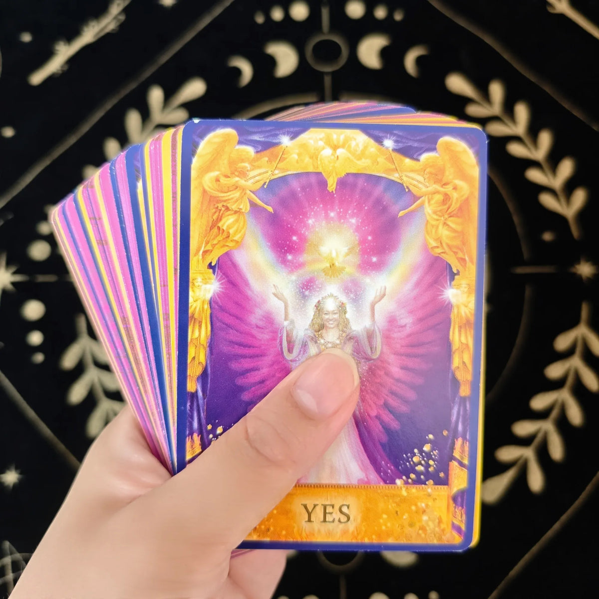 A hand holds a tarot oracle deck card showing the word YES with golden angelic imagery on a purple background.