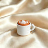 A white mug with an orange lid featuring a white heart design and a silver chain keychain, set against a silky beige backgrou