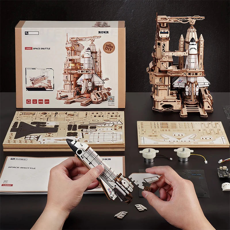 A wooden space shuttle model kit is being held by hands, displayed alongside a fully assembled model and parts.
