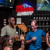A man and a woman are enjoying drinks in front of an LED neon sign featuring a beer mountain logo.