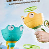 A hand is holding a yellow frog toy that launches a green flying saucer, set against a natural outdoor background.
