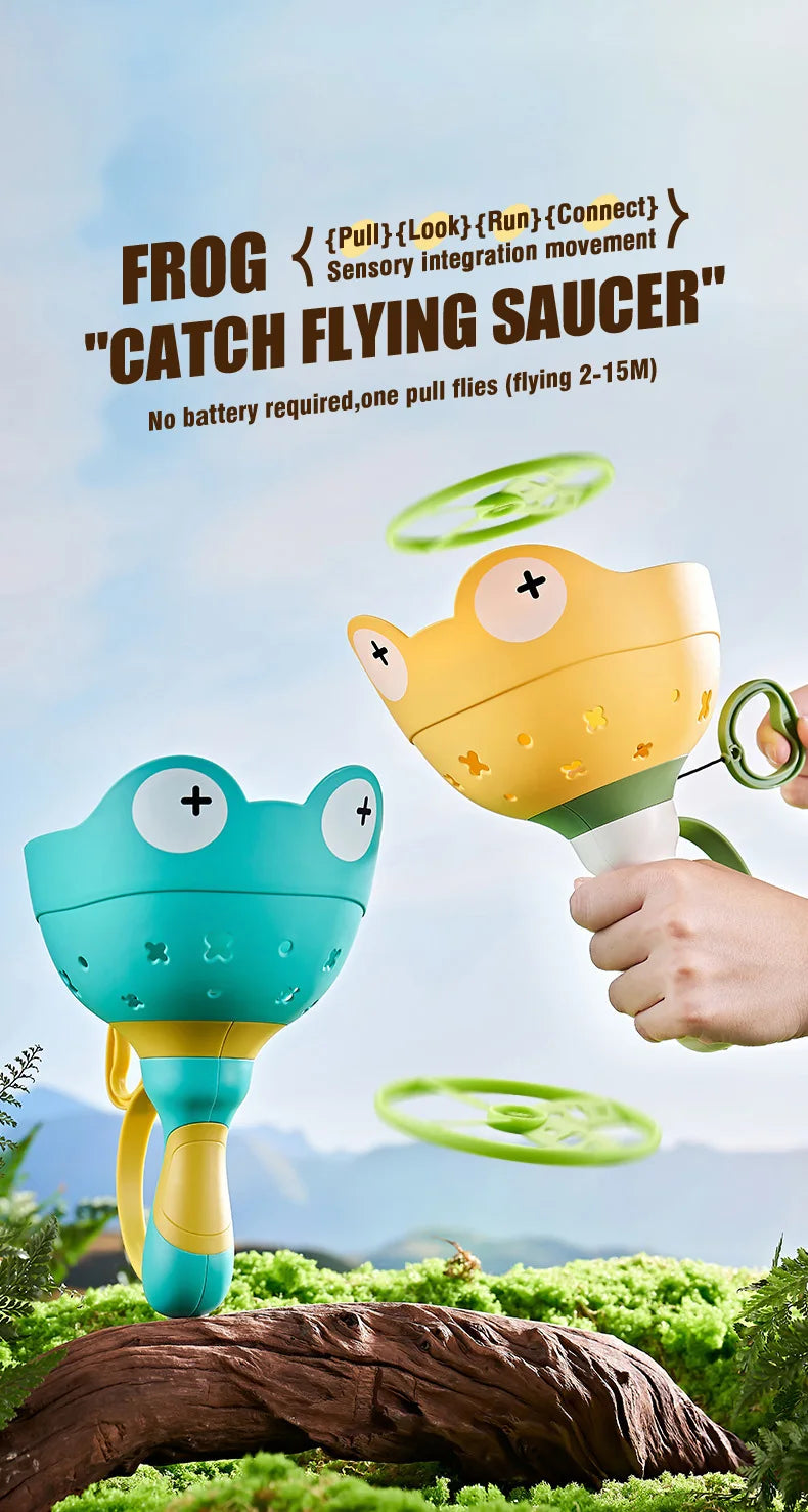A hand is holding a yellow frog toy that launches a green flying saucer, set against a natural outdoor background.