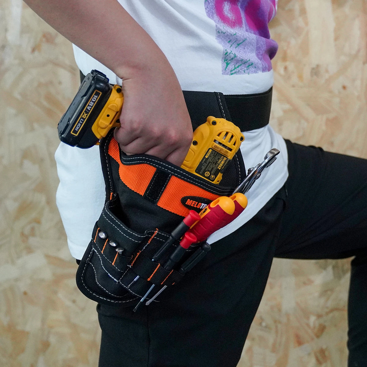 A hand holds a yellow drill tool inside an orange and black heavy duty holster attached to a belt.