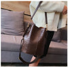 A brown alligator pattern crossbody bag is worn by a person sitting on a gray sofa.