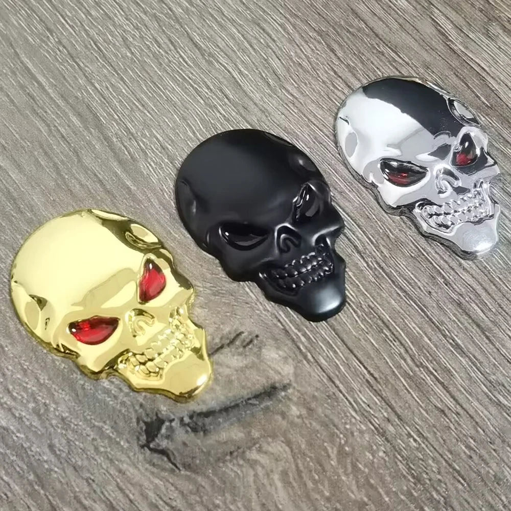Three chrome skull emblems in gold, black, and silver are displayed on a wooden surface.