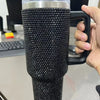 A black rhinestone bling tumbler with a clear lid, straw, and handle is held in a person's hand.