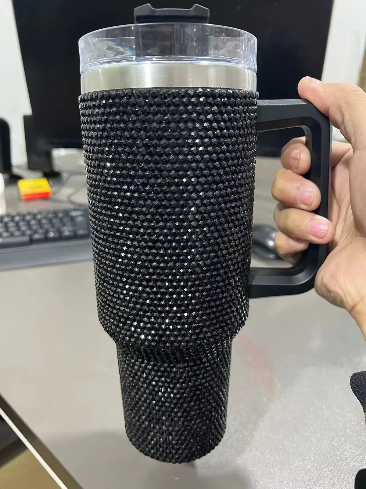 A black rhinestone bling tumbler with a clear lid, straw, and handle is held in a person's hand.