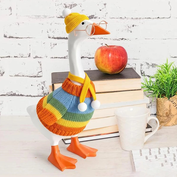 A white goose costume featuring an orange beak and yellow knitted accents, standing next to a stack of books and an apple.