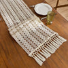 Beige bohemian farmhouse table runner with fringe detail placed on a wooden table next to a gold goblet and silverware.